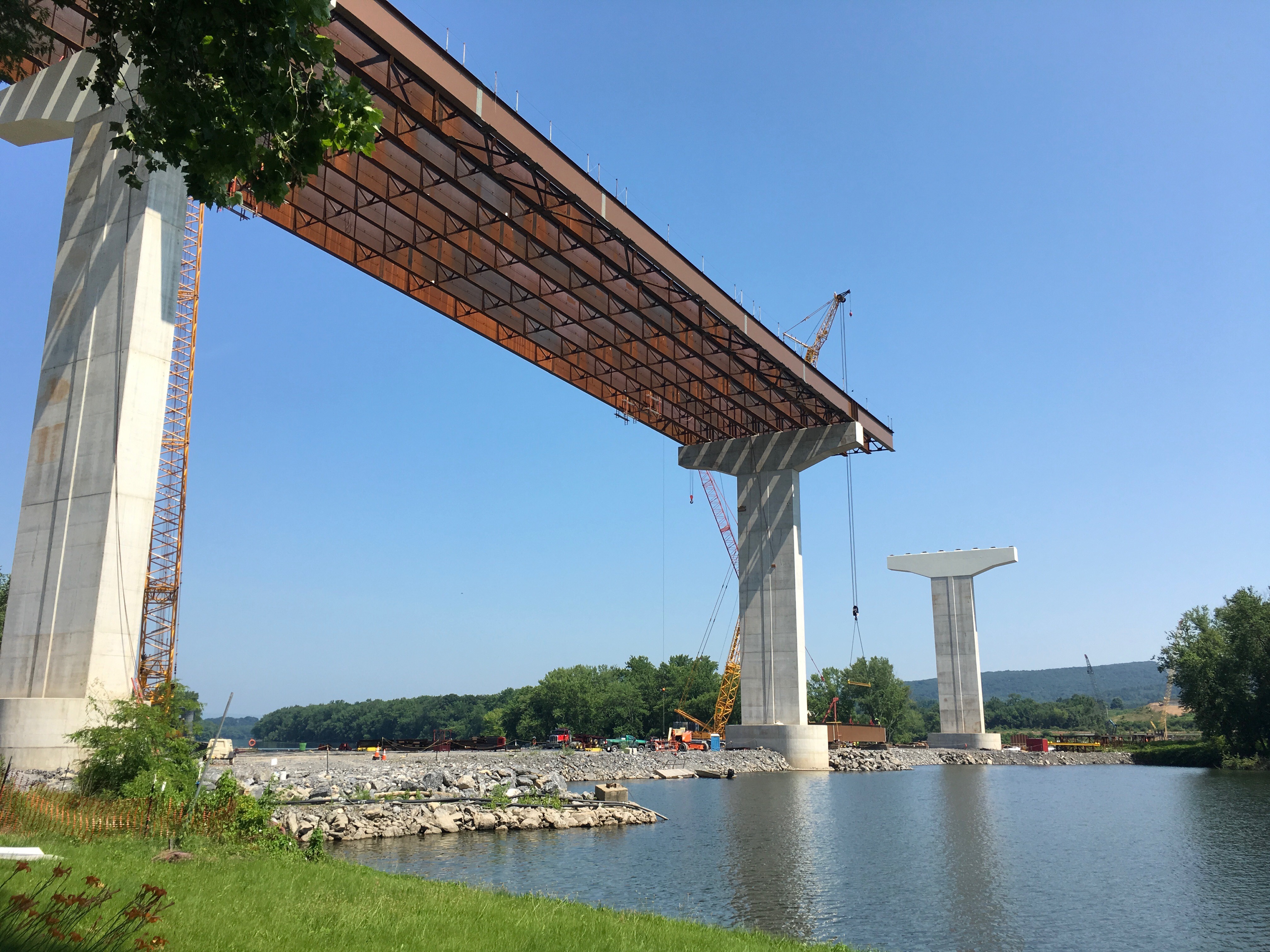 A bridge with concrete supports and steel beams is constructed along a river. In the background, a crane begins to lift a steel beam into place.